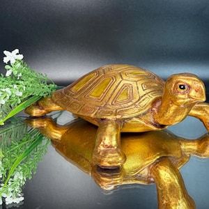 May include: A golden turtle figurine with a detailed shell pattern. The sculpture is displayed on a reflective surface, with a sprig of greenery and white flowers in the background. The turtle's shell has a geometric design.
