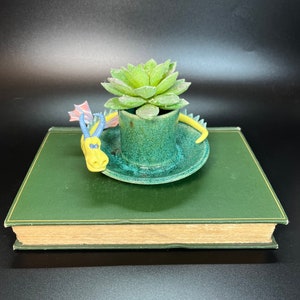 May include: A green ceramic planter shaped like a hat with a small succulent plant growing inside. A yellow and blue dragon figurine is curled around the base of the planter. The planter is sitting on top of a green book.