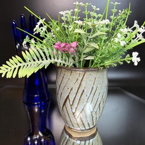 May include: A ceramic vase with a speckled light blue glaze and dark brown diagonal etched lines, filled with an arrangement of green foliage, white flowers, and a small cluster of pink blossoms, positioned in front of a dark blue glass hand form.