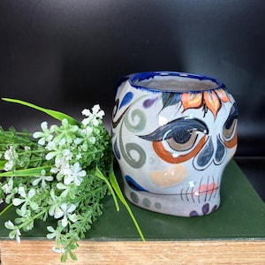 May include: A ceramic skull-shaped container with a blue rim and intricate painted details. The design features floral and swirl patterns in orange, blue, and green on a light gray base. A sprig of white flowers and green leaves rests beside the container.