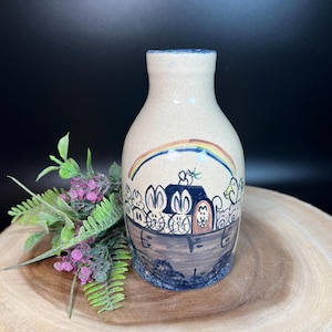 May include: A beige ceramic vase with a blue rim and base. The vase features a hand-painted illustration of Noah's Ark with animals and a rainbow. The vase is approximately 6 inches tall.