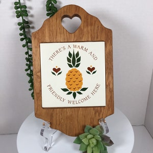 May include: A wooden sign with a heart-shaped cutout at the top. The sign has a white tile with a pineapple design and the text "There's a warm and friendly welcome here".