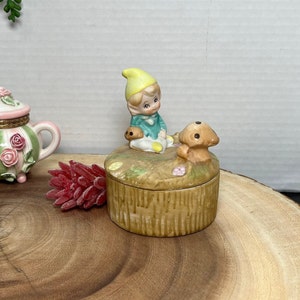 May include: A ceramic trinket box with a whimsical design featuring a small elf sitting with a squirrel. The box is decorated with a brown, green, and yellow color scheme and has a textured surface.