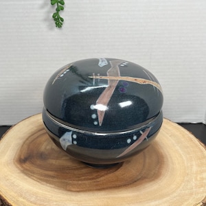 May include: A dark blue ceramic bowl with a lid, featuring abstract patterns in brown, white, and black. The bowl is sitting on a wooden base.