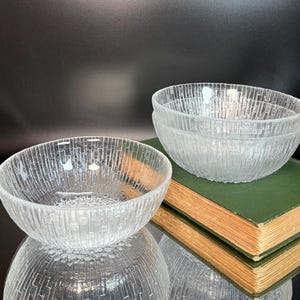 May include: Clear glass bowls with a textured, vertical lined pattern. One bowl is in the foreground, with two more stacked on a green book in the background. The bowls have a simple, yet elegant design.
