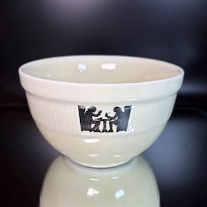 May include: A cream-colored ceramic bowl with a ribbed exterior texture. The front of the bowl features a black graphic illustration of two figures sitting at a small table, each holding a pipe, set against a smooth, plain background surface.