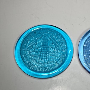 May include: Two round, blue glass objects with a lighthouse design. The objects have a raised relief design with the text "USS Houston" and "1938". The glass is a vibrant, transparent blue.