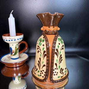 May include: A ceramic bell-shaped figure with a brown hat and a cream-colored body decorated with green and brown floral patterns, positioned next to a ceramic candle holder with a white candle and the text Stratford-upon-Avon written on it.
