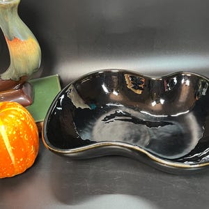 May include: A black ceramic serving bowl with a wavy, organic shape. The bowl has a glossy finish and a subtle brown rim.