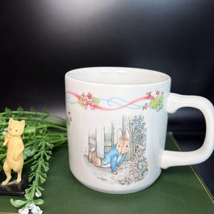 May include: White ceramic mug featuring a Peter Rabbit illustration. The mug has a curved handle and is decorated with a floral and ribbon design. A small figurine of a cat is also present.