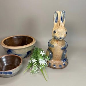 May include: A ceramic rabbit figurine with a tan and blue speckled glaze, accompanied by two matching ceramic bowls with blue painted accents and a small bunch of artificial white flowers with green stems and leaves.