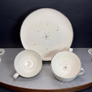 Puede incluir: Un plato de cerámica color crema y dos tazas de té a juego con un borde plateado y un delicado patrón de estallido de estrellas en gris y dorado. El plato está colocado detrás de las dos tazas, cada una con un asa y un diseño de estrella.