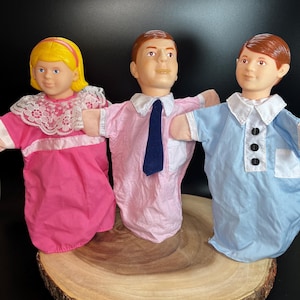 May include: Three hand puppets, one female with blonde hair wearing a pink dress with white lace trim, and two males with brown hair wearing pink and blue shirts with collars and ties.