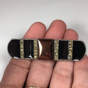 May include: A black and silver brooch with a central square of rose gold. The brooch is decorated with rows of clear rhinestones.