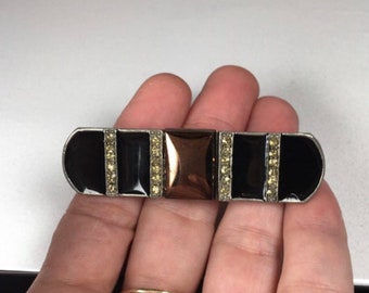 May include: A black and silver brooch with a central square of rose gold. The brooch is decorated with rows of clear rhinestones.