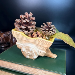 May include: A decorative ceramic cart planter with a yellow interior, filled with pine cones and a leaf. The cart is light brown with carved wood details and white accents on the wheels. The background is dark.