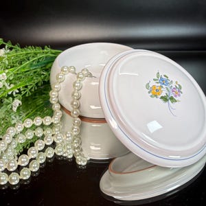 May include: White ceramic lidded dish with a floral design and a pearl necklace. The lid features a bouquet of blue, yellow, and purple flowers. The dish has a light pink and blue border. Greenery is visible in the background.