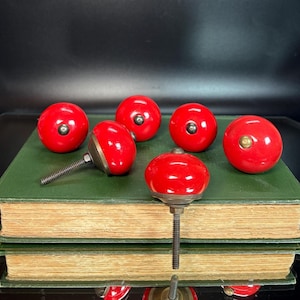 May include: Six bright red cabinet knobs with metal hardware are arranged on a stack of green books. The knobs are round with a glossy finish, and the metal hardware has a silver and gold tone.
