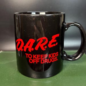 Pode incluir: Uma caneca de cerâmica preta com o texto vermelho D.A.R.E. em uma fonte negrito e estilizada, seguido pelo texto vermelho TO KEEP KIDS OFF DRUGS. A caneca tem um acabamento liso e brilhante e uma alça curva no lado direito da estrutura.