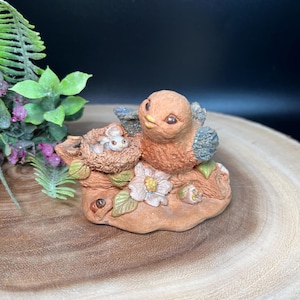 Op de afbeelding: Gedetailleerd beeldje van een vogel met blauwe vleugels en een nest met een babyvogel. Het beeldje is gemaakt van een terracotta-kleurig materiaal en heeft een witte bloem en groene bladeren.