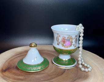 Könnte beinhalten: Eine weiße Porzellan-Schmuckdose mit grünem und goldenem Rand. Der Deckel hat einen goldenen Knopf und ein Blumenmuster. Die Dose steht auf einer Holzoberfläche mit einer Perlenkette im Inneren.