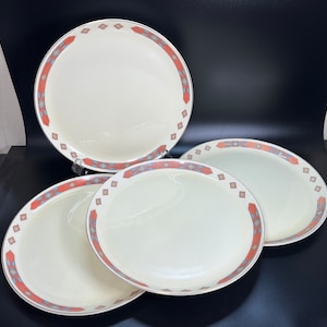 May include: Four white ceramic dinner plates with a red, blue, and gold geometric pattern around the rim.