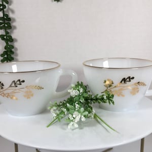 Puede incluir: Dos tazas de té blancas con diseños florales dorados y bordes dorados. Las tazas están sobre un platillo blanco.