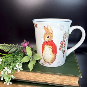 May include: White ceramic mug featuring a charming illustration of a brown rabbit in a red jacket. The mug has a blue rim and a white handle. Floral and butterfly accents decorate the mug. The mug is sitting on a stack of green books.