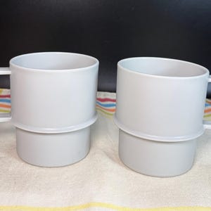 May include: Two light gray mugs with a smooth, cylindrical body and a distinct horizontal ridge around the center. Each mug features a square-shaped handle attached to the side, providing a clean, uniform appearance for these beverage containers.