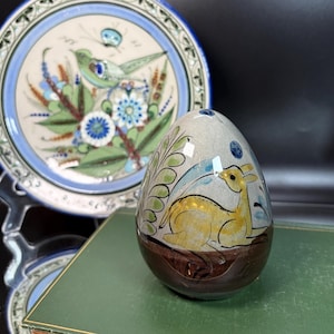 May include: A ceramic egg-shaped decorative item with a deer design in yellow and brown, set against a light gray background. The egg is adorned with blue and green accents. A decorative plate with a bird and floral design is in the background.