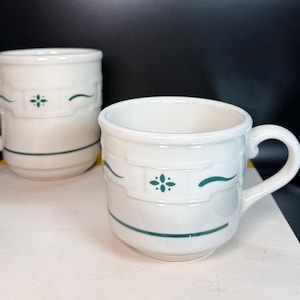May include: Two white ceramic mugs with green accents. Each mug has a handle and a decorative design featuring a green floral pattern and curved lines. The mugs are on a white surface.