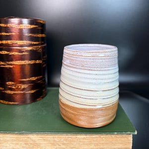 May include: A handmade ceramic tumbler with a textured, layered design. The cup features a light brown base, transitioning to white and light blue hues towards the top. A dark brown container is visible in the background. The objects are on a green book.
