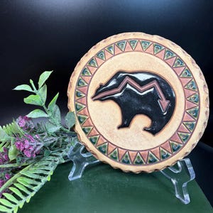 Puede incluir: Un posavasos redondo de cerámica de color beige con un diseño de oso negro y un rayo rojo y blanco. El posavasos tiene un borde decorativo con triángulos verdes y marrones.