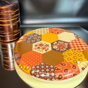 Peut inclure: Un récipient rond vintage avec un couvercle et un motif patchwork dans les tons orange, marron et jaune. À côté, un récipient cylindrique et haut avec un aspect brun foncé et stratifié. Les deux objets sont sur une surface réfléchissante.