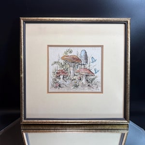 Pode incluir: Uma ilustração emoldurada apresentando um grupo de vários cogumelos em tons de castanho, vermelho e branco, acompanhados por pequenas flores azuis e folhagem verde. A obra está colocada dentro de um passe-partout creme e uma moldura dourada texturizada.