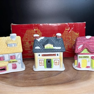 Op de afbeelding: Drie keramische miniatuurhuisjes met besneeuwde daken en gedetailleerde gevels. De huisjes zijn gelabeld met "The Baker", "The Queen's Inn" en "The Grocer". Een rode doos met huisillustraties staat op de achtergrond. De huisjes staan op een houten oppervlak.