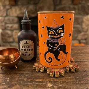 May include: A cylindrical orange canister with a bamboo lid featuring a black cat illustration wearing a party hat and holding a pumpkin lollipop, surrounded by black stars. Next to it is a dark glass bottle labeled FEAR, WRATH & MAGIC.