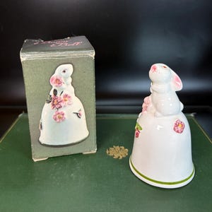 May include: A white ceramic bunny bell with pink floral accents and a green rim. The bell features a bunny figurine on top and comes with its original box. The box has a matching design of the bell.