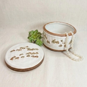 May include: A round ceramic lidded jar with a white glaze and brown rim, featuring a repeating pattern of small brown trees. A string of white pearls rests inside the open jar, and a small green succulent sits beside the circular lid.
