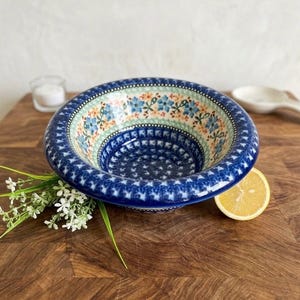 May include: A round ceramic bowl featuring a wide, dark blue rim with a white star pattern. The interior displays a band of blue flowers and orange accents on a light green background, with a deep blue center decorated with small white star motifs.