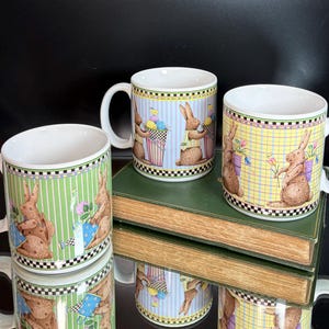 May include: Three ceramic mugs with Easter bunny designs. Each mug features a white handle and a rim with a black and white checkered pattern. The mugs have different background colors: green stripes, blue stripes, and yellow plaid. The bunnies are holding baskets and flowers.