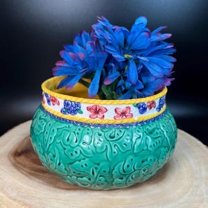 Könnte beinhalten: Ein grüner Keramiktopf mit gelbem Rand und floralem Design. Der Topf hat ein erhabenes Blattmotiv und ist mit rosa und blauen Blumen verziert. Ein Strauß blauer Blumen steht im Topf.