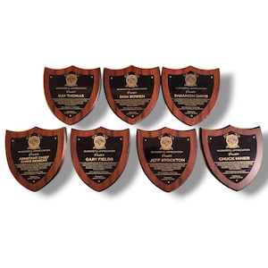 May include: Seven wooden shield-shaped plaques with dark brown frames and black centers. Each plaque has gold text and a gold emblem. The plaques are arranged in a semi-circle. The text includes names and the phrase "In Grateful Appreciation".