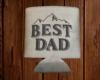 May include: A white beverage holder with the words "BEST DAD" in dark gray block letters. Above the text is a mountain range graphic. The holder is set against a wooden background.
