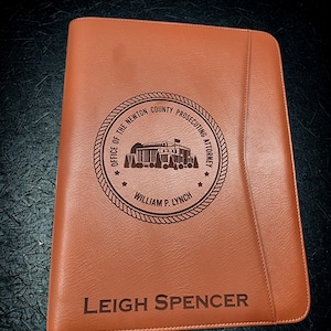 May include: A brown leather portfolio with the text "LEIGH SPENCER" at the bottom. The top of the portfolio has a circular emblem with the text "OFFICE OF THE NEWTON COUNTY PROSECUTING ATTORNEY" and "WILLIAM P. LYNCH".