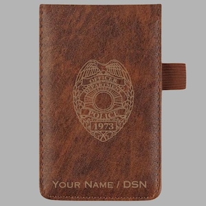 May include: A rectangular brown faux leather notebook cover with a textured finish. The front features an engraved police badge emblem with the text OFFICER DEPARTMENT POLICE 1973. Below the emblem, the text YOUR NAME / DSN is engraved. A brown elastic pen loop is attached to the right side.