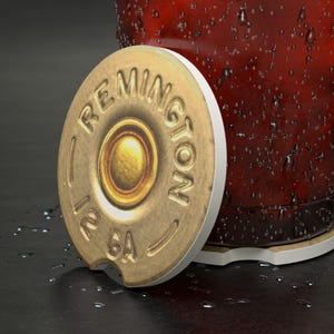 May include: A round coaster with a design resembling a 12-gauge Remington bullet. The coaster is gold with white trim and features the text "Remington" and "12 GA". The coaster is next to a glass with a red liquid.