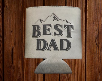 May include: A white beverage can cooler with the words "BEST DAD" in dark gray block letters. Above the text is a mountain range graphic. The cooler is set against a wooden background.