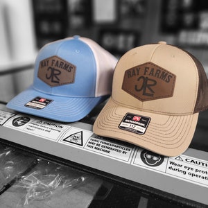 May include: Two baseball caps with brown leather patches that say "Ray Farms JR" in black lettering. The caps are brown and blue with white mesh backs.
