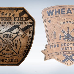 May include: Two wooden plaques with black engraved text. The left plaque is shaped like a shield and features the text "Exeter Fire Protection District", "Fire", "Rescue", "Jim Matthew", and "1967-2021". The right plaque is shaped like a shield and features the text "Wheaton Fire Protection District", "Fire/Rescue", and "Established 2013".
